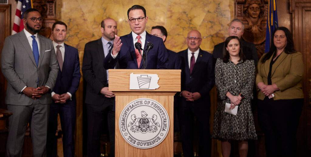 PA Governor Josh Shapiro stands behind a podium, while a group of people stand behind him.