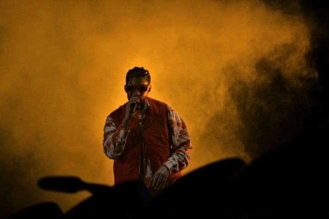 Rapper Bad Bunny, wearing a red and white jacket and sunglasses, sings into a microphone against a orange smoke background.