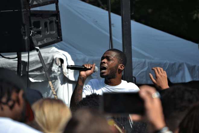 Philadelphia rapper Kur, a Black man wearing a white t-shirt, performs into a cordless microphone.