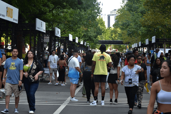 People walk along a street that's been closed to vehicular traffic, between trees and signage for Cause Village at Made in America 2022.