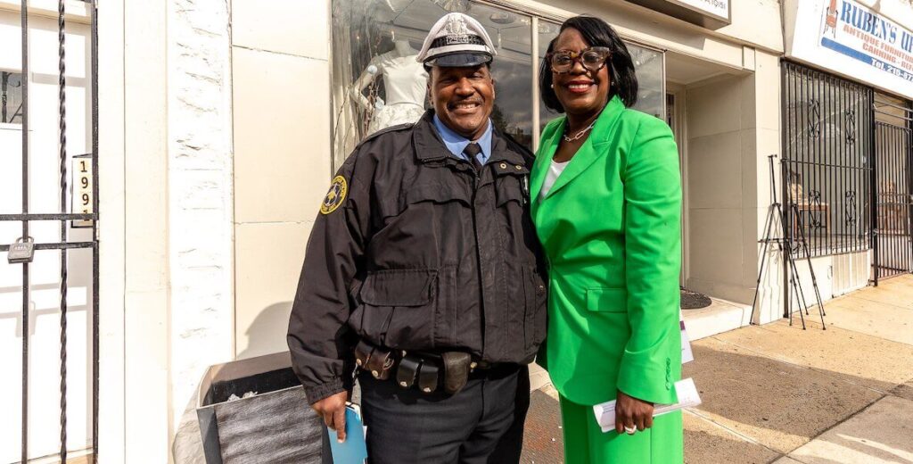 Presumptive Mayor of Philadelphia Cherelle Parker, a Black woman, wears a bright green pantsuit and stands next to a Philadelphia police officer on a sidewalk. This is for a story about public safety.