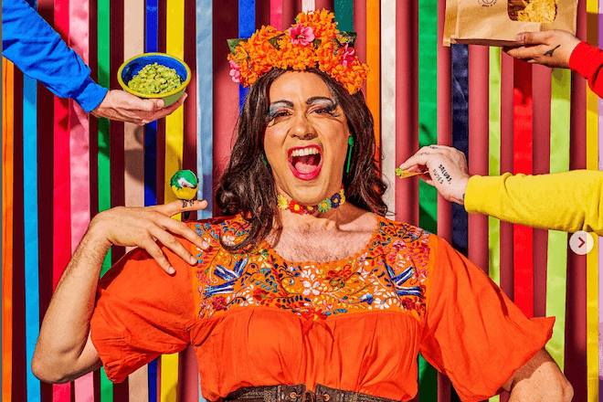Drag performer Martha Graham Cracker poses against a colorful striped backdrop for the cover of Mission Taqueria's ¡Saluds Dudes! calendar. Reaching toward her are hands holding tortilla chips, guacamole ... 