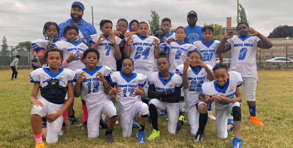 The South Philly Sharks youth football players pose for a team photo in white uniforms with blue numbers and their coaches standing behind them