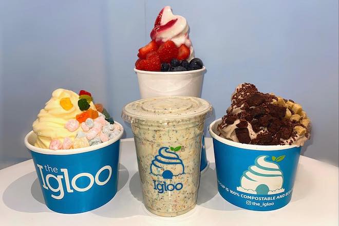 Soft-serve desserts: a yellow soft serve with mochi, a white and red soft serve topped with strawberries and blueberries, a multi-colored milkshake, and a loaded cup of chocolate soft serve and toppings stand on a table, all in "Igloo" cups.