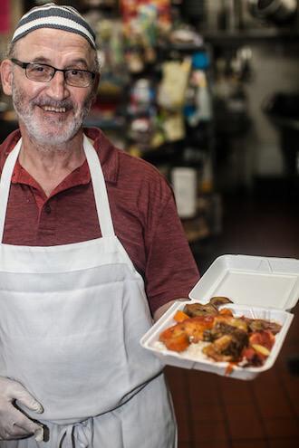 A man with a grey-and-white bear and glasses wearing a white apron and red polo shirt and a small striped hat smiles while holding a plate of shawarma from Al Amana in Philadelphia.