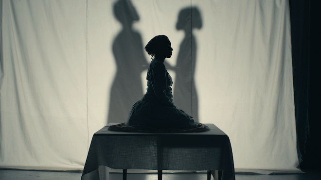A silhouette-looking still from BlackStar Film Festival documentary short "Bone Black: Midwives vs. the South" directed by Imani Nikyah Dennison. On it, an African American woman wearing a headscarf appears to be melting into a table. Behind her: Projected on a white sheet behind her: shadows of a woman and a girl.