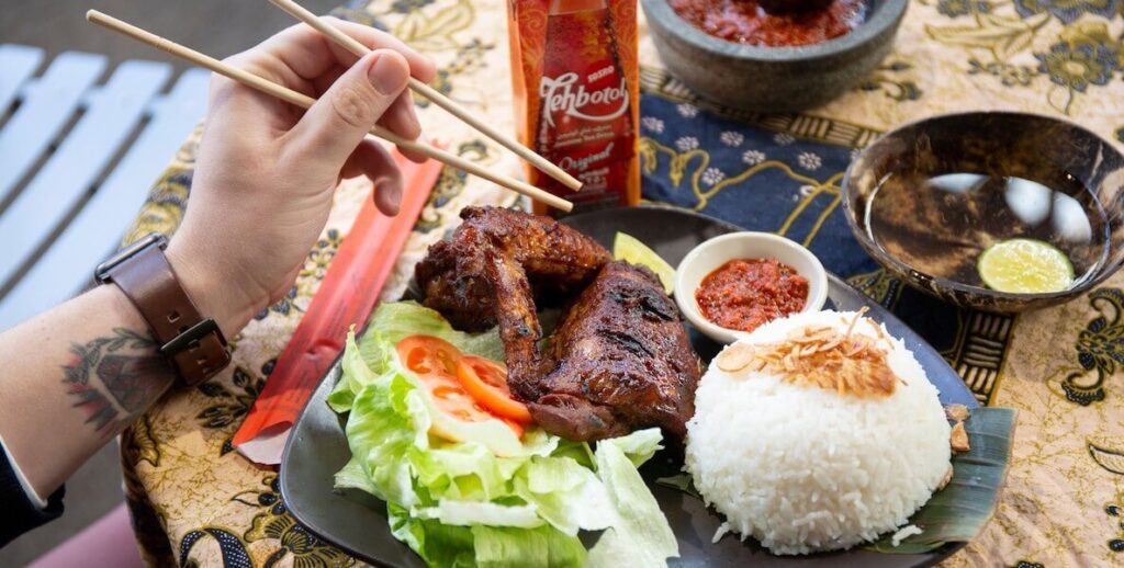 A hand holding chopsticks reaches toward a plate of meat, lettuce, and rice from Hardena, an Indonesian restaurant in Philadellphia.