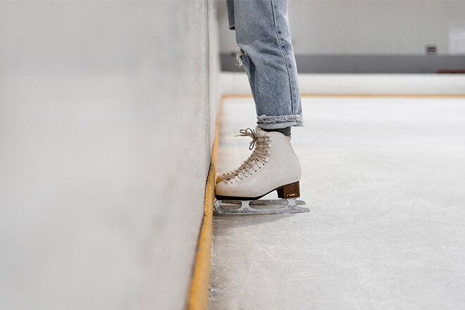 Partial view of legs and feet of a person in jeans and white ice skates next to the edge of an ice rink.