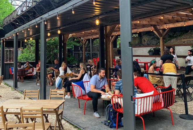 People sit on red benches on a covered platform beneath a rail bridge at Love City Brewing Co's "Gardens" in Philadelphia.