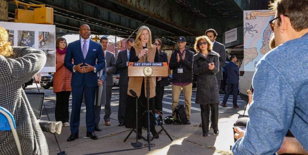NYC Sanitation Commissioner Jessica Tisch, a blonde woman with a dark suit and brown vest stands at a podium beneath an overpass, and speaks to a crowd. To her right: NYC Mayor Eric Adams. Courtesy of the New York City Department of Sanitation.