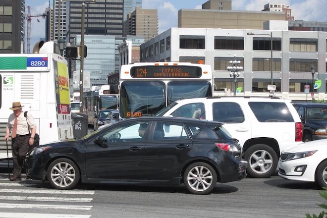 A man in a hat and tie walks with a cane across an intersection crowded with cars and SEPTA buses in Philadelphia.