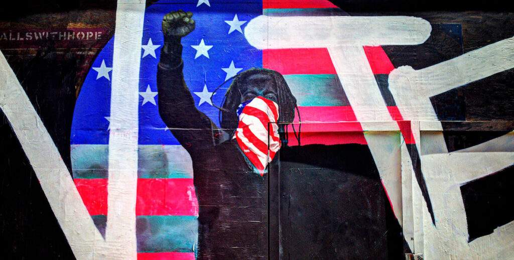 A mural shows a man painted mostly in black with mid-length hair and a red-and-white-striped banana over his mouth raising his right fist. Backdropping him in white are the letters V, T and E. Between the V and T is a circle (an O) with the U.S. flag.