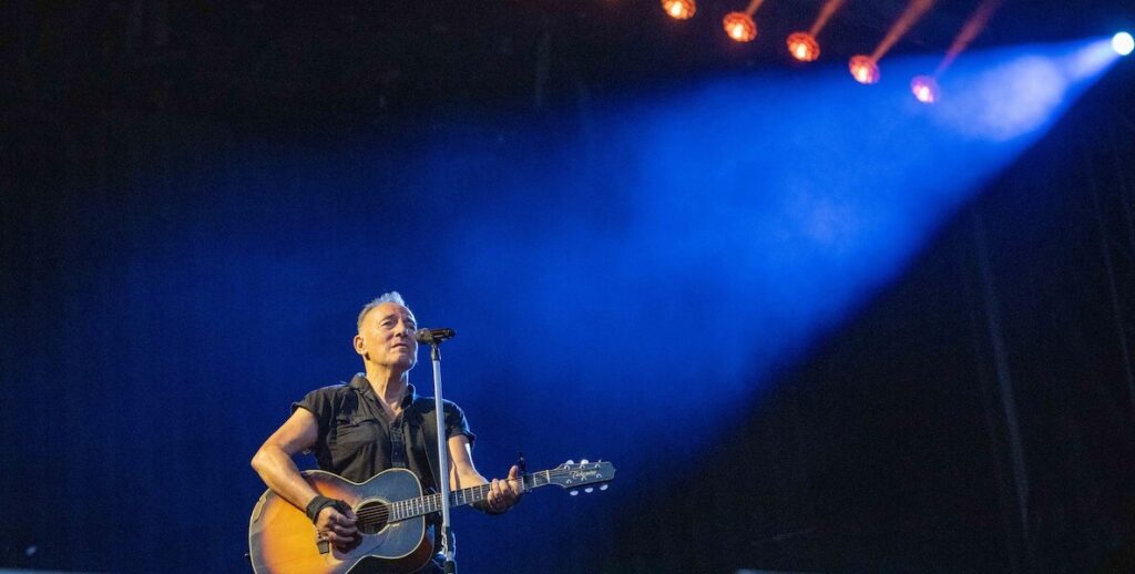 Bruce Springsteen stands in a spotlight, facing the camera and an unseen audience, holding his guitar and standing in front of a microphone in 2023.