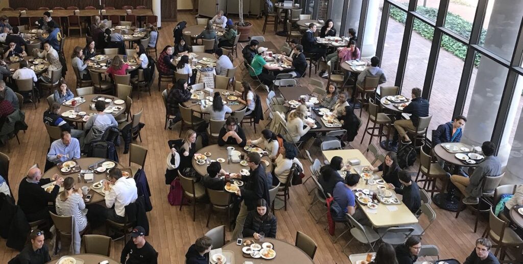 Students sit and eat at tables in a large dining hall. This story is about hunger among college students, and how we can remedy it.