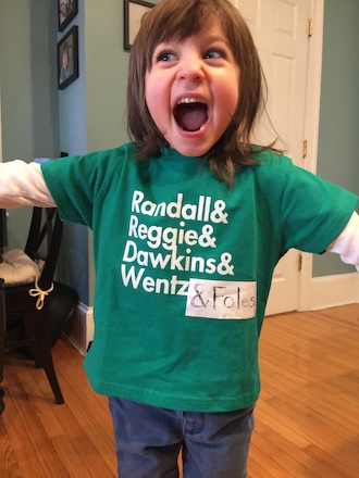 A young white girl with brown hair (bangs, shoulder-length), smiles with her mouth wide open. She is wearing a green t-shirt that reads "Randall & Reggie & Dawkins & Wentz," with an added piece of white tape that reads "& Foles."