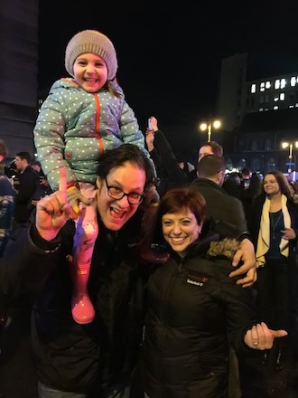 A small child in a winter coat and hat sits on the shoulders of a white man with glasses and a black jacket, standing alongside a smaller white women in a black coat on Broad Street following Super Bowl LII.