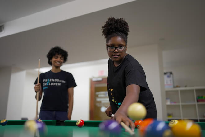 Two people wearing black t-shirts play pool. One, a woman with black hair piled high on her head and glasses, aims the cue at the balls. They are participants in Friends of the Children in Boston.