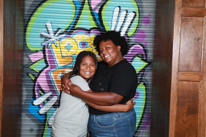 A girl and a young woman hug in front of a graffiti artwork. They are participants in Friends of the Children in Detroit, Michigan.