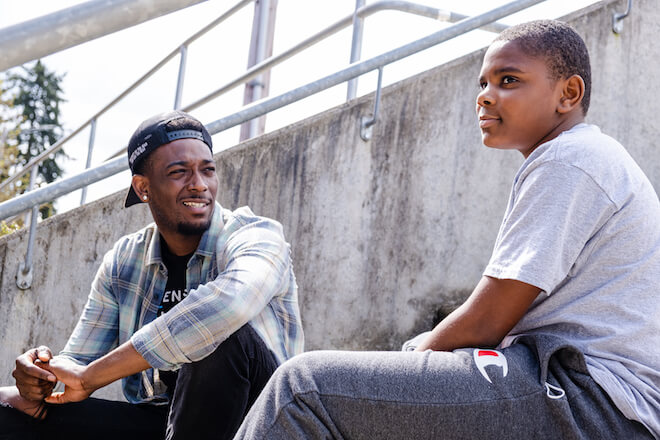 A Black man in a plaid flannel shirt, black jeans, a backwards cap with a diamond earring sits in a stadium alongside a Black book with closely cropped hair and wearing a t-shirt and sweatpants. They are participants in the Portland, Oregon-based Friends of the Children program.