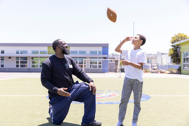 A bearded Black man in sweats takes a knee on a football field alongside a Black boy in sweatpants and a white t-shirt who is tossing a football in the air. They are participants in Friends of the Children in San Francisco, CA. A school building is in the background.