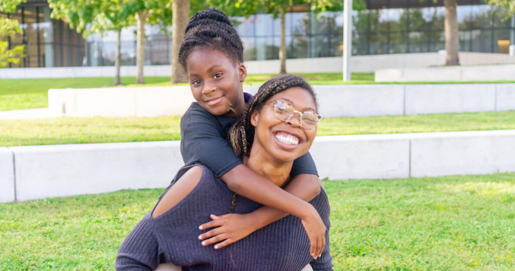 A young Black girl in a black top rides on the back of an older Black woman, also in a black top, and with braids and glasses. They both smile. They art part of the nonprofit organization Friends of The Children in Tampa Bay, FL.