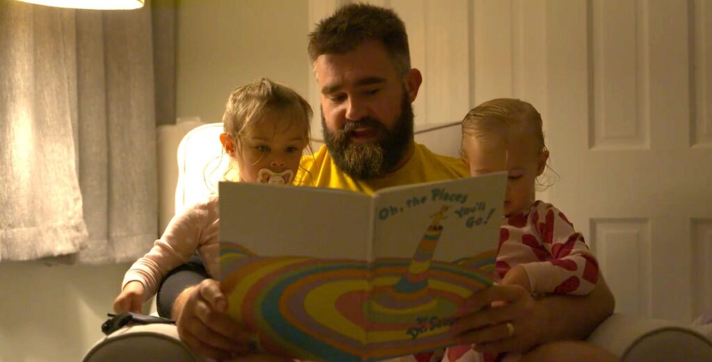 A large White man with a beard (Eagles center Jason Kelce) sits in a chair reading the Dr. Seuss book "Oh, the Places You'll Go" to his two toddler daughters, sitting on his lap.