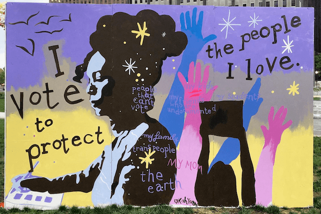 A colorful mural features a profile of a painter, hair piled high on their head, dipping a brush into a paint set. Behind the painter are silhouettes of raised hands and a person holding a sign The mural is for Power to the Polls, and was painted by Kah Yangni. It reads, "I vote to protect the people I love."