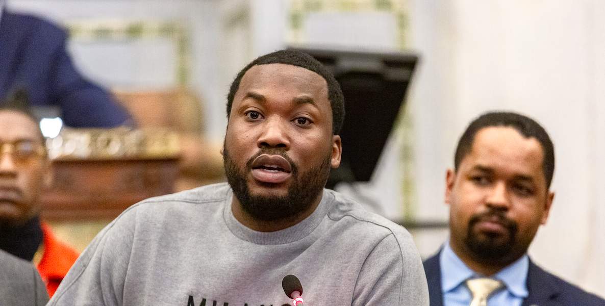 Robert “Meek Mill” Williams wears a grey sweatshirt and speaks into a microphone at a 2019 meeting of Philadelphia City Council. Behind Meek Mill sits Senator Sharif Street in a suit and tie.