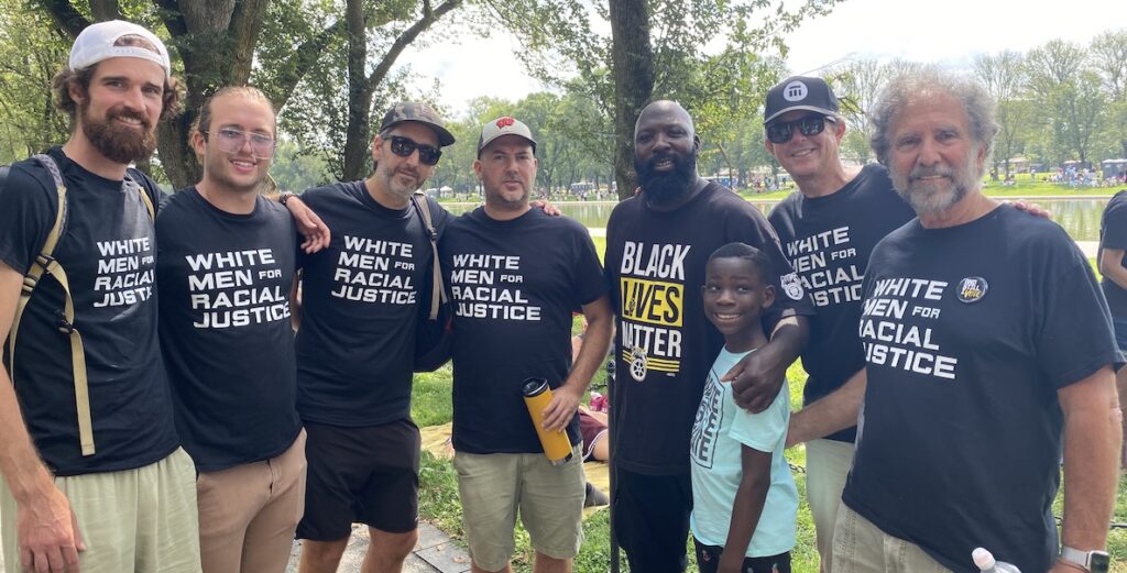 Six White men wearing White Men for Racial Justice t-shirts, posing with a Black man in a Black Lives Matter t-shirt and his son.