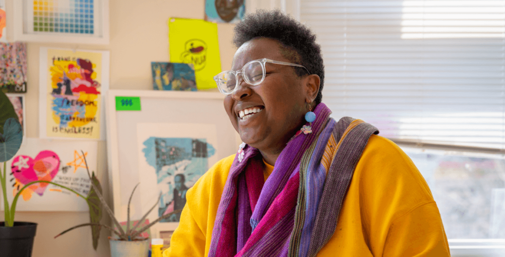 Artist Kah Yangni smiles in their Philadelphia studio. They are African American, with short hair and clear glasses, wearing a yellow sweater layered with purple scarves.