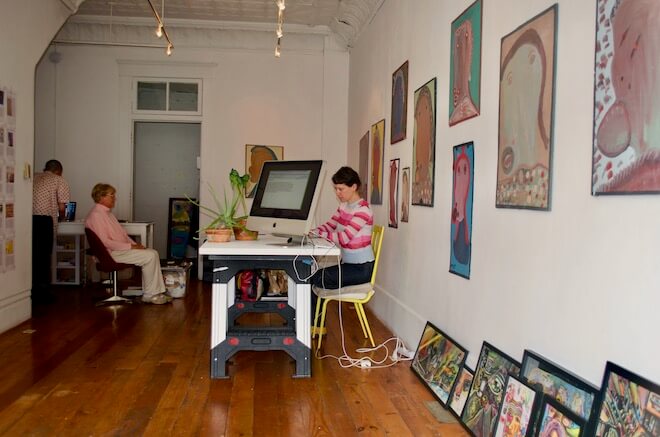 Art critic Lori Waxman, a white woman with short brown hair wearing a striped sweater, sits at a desk typing in an art gallery, with paintings and artwork behind her. Facing away from her, a monitor displays what she's typing.