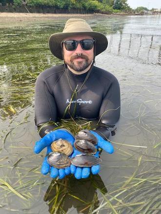 Matt Gentry, a bearded white man wearing a black wetsuit, a beige hat, sunglasses, a nosering and blue latex gloves, stands in a marsh, presenting five separate kinds of freshwater mussels in his hands.