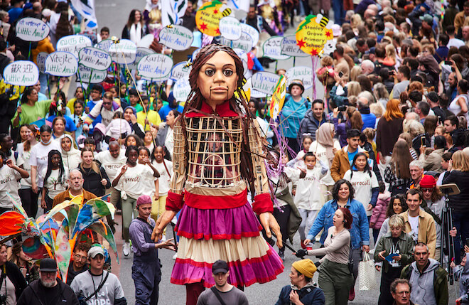Little Amal, a giant puppet representing a 10-year-old Syrian refugee, walks through a crowd of people carrying signs.