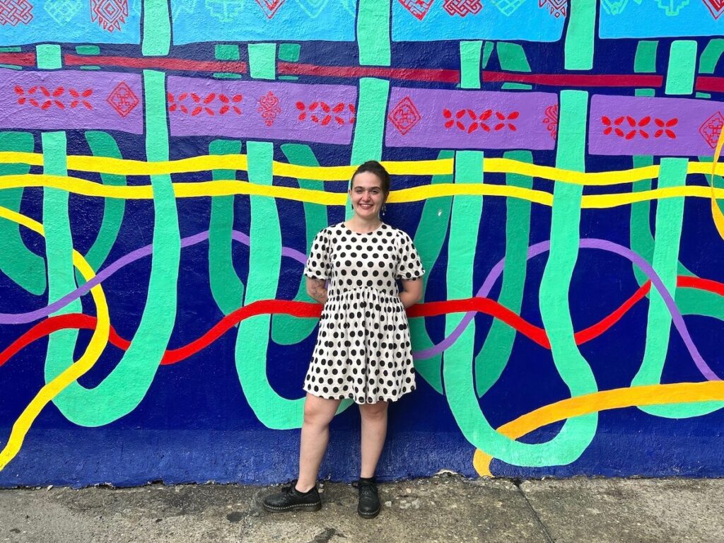 A white woman in a polka dot dress stands in front of a colorful mural depicting threads. This is Isa Segalovich, a social media art commenter.