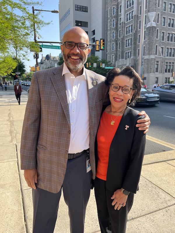 Two people stand outside on a sidewalk: A tall black man with a bald head, glasses and beard, and a petite Black woman with glasses, a suit jacket and a red blouse. They are Bret Perkins and JoAnne Epps