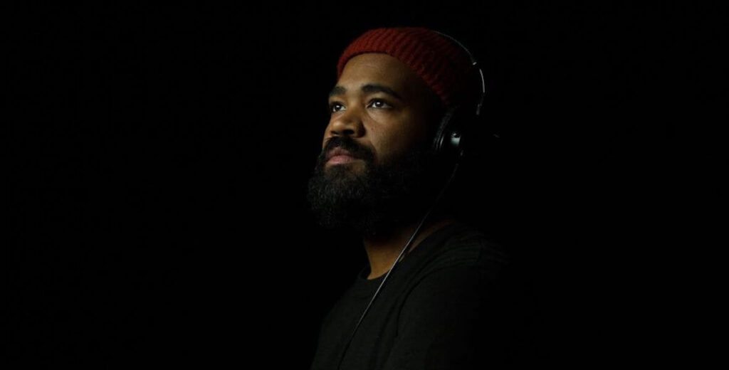 Artist, VJ and DJ Rashid Zakat, a Black man with a short beard wearing headphones, a red beanie and a black shirt, seems to blend into the background in this portrait.