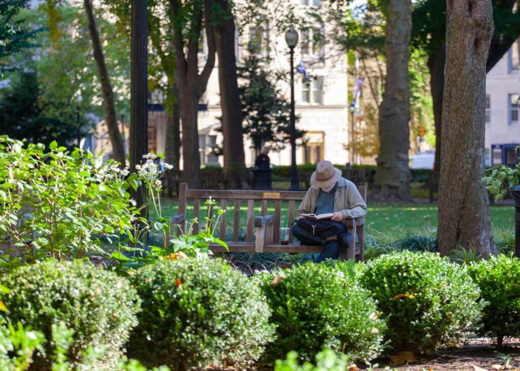 A white man in a beige hat and jacket reads a book on a bench in Rittenhouse Square in Philadelphia.