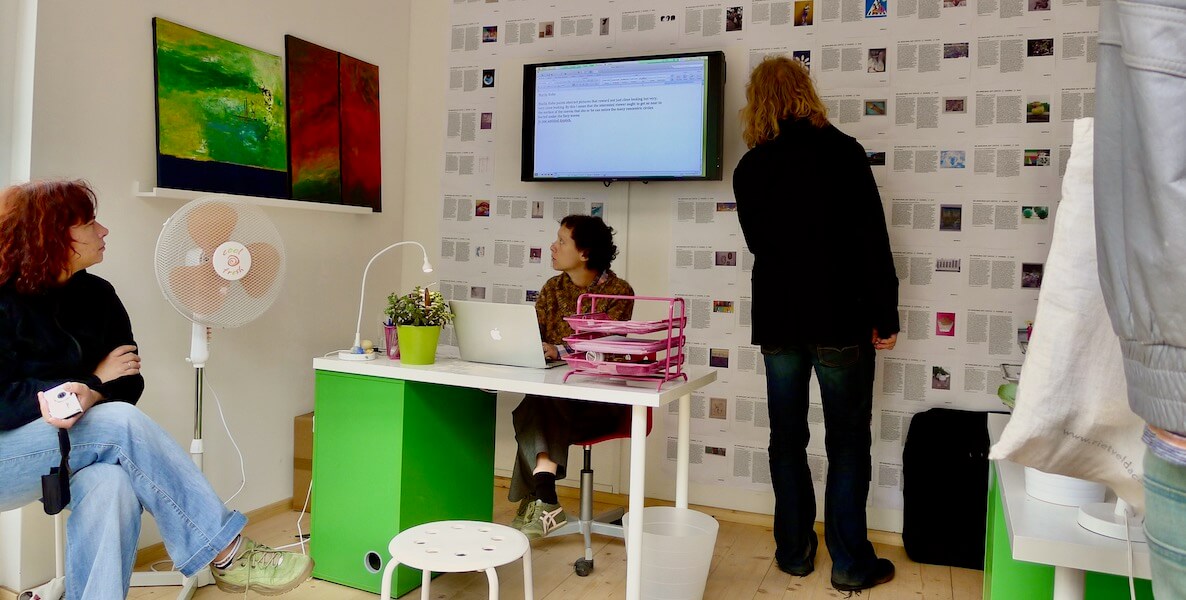 Three women in a small gallery space. In the middle is art critic Lori Waxman, a white woman with short brown hair. She sits behind a white and green desk, typing on an Apple laptop while looking at two paintings on a shelf to her right. Behind her is a wall of typed-up reviews alongside small renderings of artwork and a monitor.