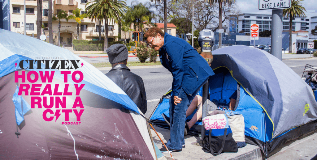 Los Angeles Mayor Karen Bass, an African American woman in a blue coat, blue jeans, and eyeglasses stands between two tents pitched next to a street in Los Angeles, speaking to a person in a black jacket and hoodie who appears to be living there. Karen Bass is a guest in The Philadelphia Citizen's "How to Really Run a City" podcast.