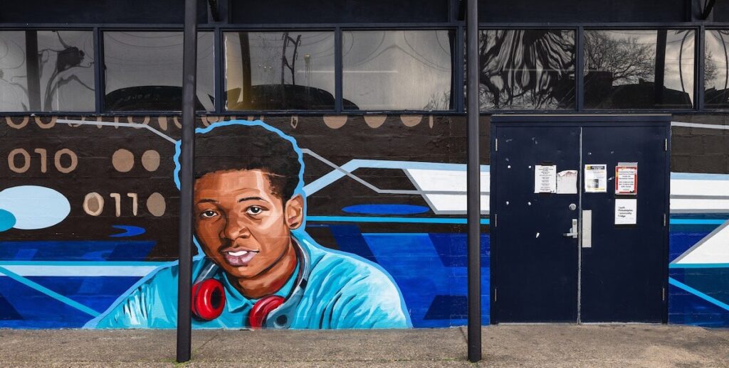 On the exterior wall of a Philadelphia recreation center is a mural of a Black boy wearing a blue shirt, with red headphones on his neck.