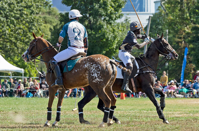Polo players on horseback play on a field with the Philadelphia skyline in the background.