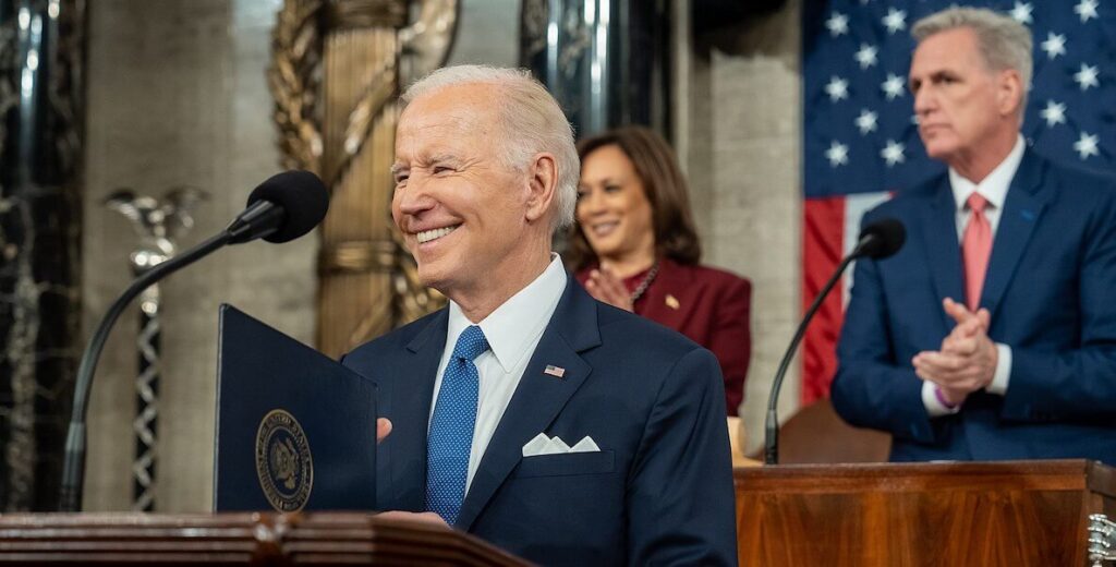 President Joe Biden stands at a podium to deliver the 2023 State of the Union. Behind him, Vice President Kamala Harris and Speaker of the House Kevin McCarthy applaud. Behind them hangs the U.S. flag.