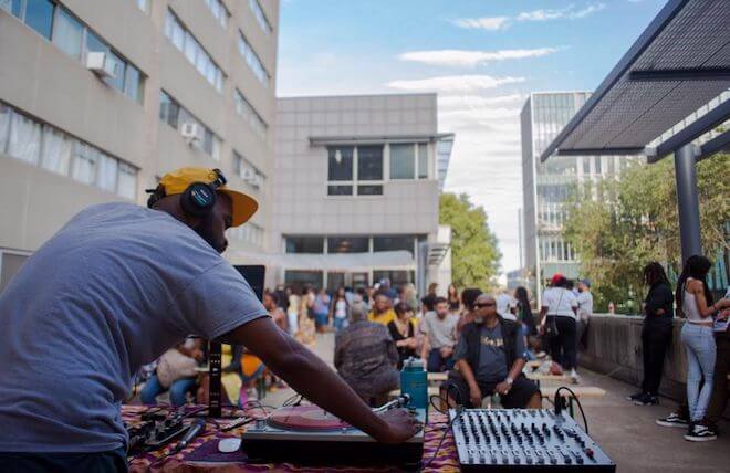 DJ, VJ and artist Rashid Zakat, a Black man wearing a grey t-shirt, yellow cap and headphones, works a turntable at an outdoor event in Philadelphia.