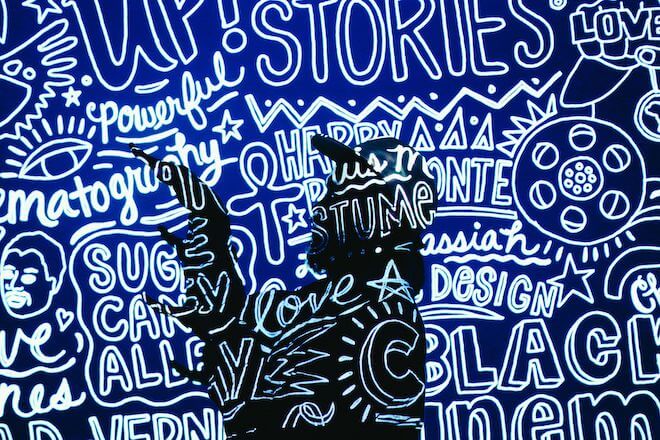Words, mostly bubble letters and some cursive combine with a few sketches in white on a blue background. In the middle is the silhouette of a man with a beard wearing a baseball cap. This is artist Rashid Zakat of Philadelphia.