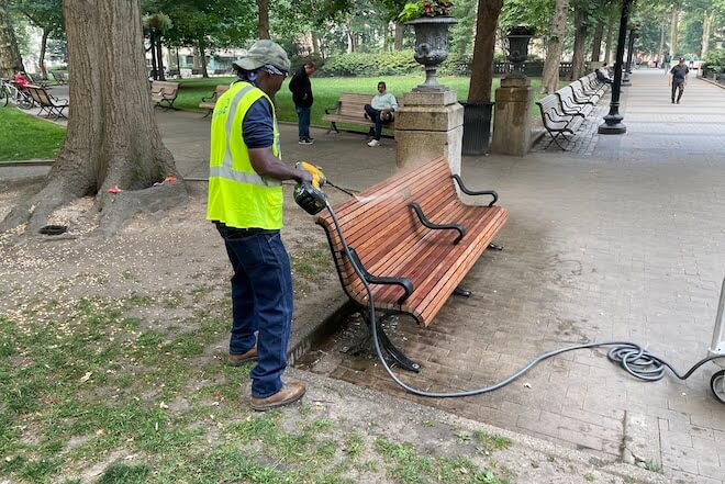 A person in blue jeans and a neon yellow vest hoses off a park bench in Rittenhouse Square.