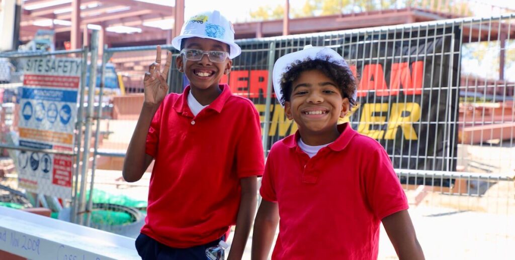 Two students wearing red polo shirts and hard hats smile while standing in front of a construction site in Philadelphia.