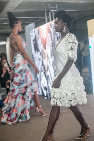 Models in floral dresses, one colorful and knee-length, and one all white and knee-length, walks the runway in a parking garage during Philadelphia Fashion Week.