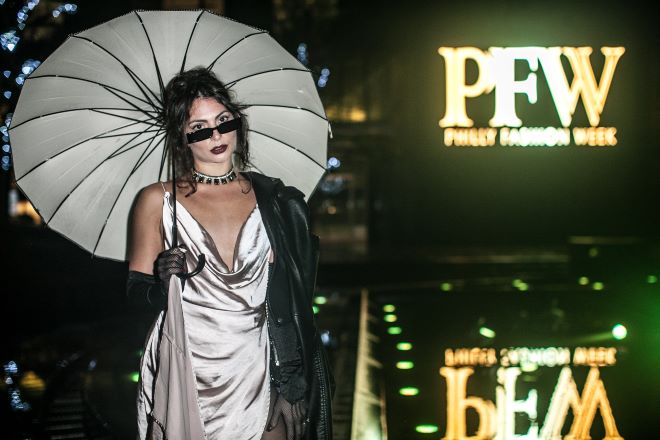 A model in a metallic Naomi Campbell dress carrying an umbrella and wearing sunglasses and sheer black gloves stands by the PFW logo for Philadelphia Fashion Week.