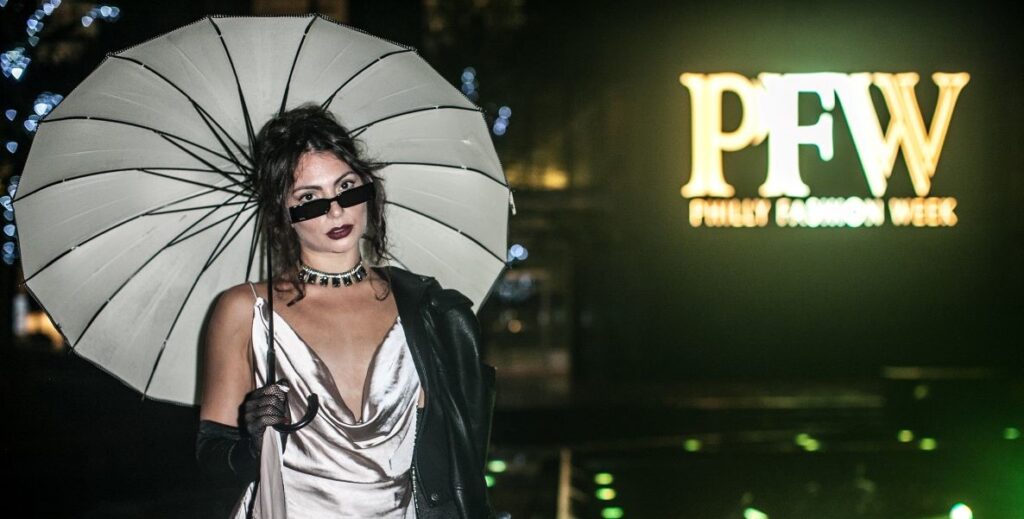 A model in a metallic Naomi Campbell dress carrying an umbrella and wearing sunglasses and sheer black gloves stands by the PFW logo for Philadelphia Fashion Week.