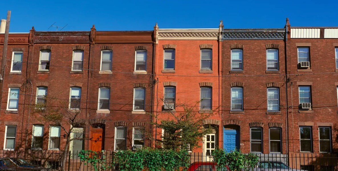 Five brick rowhomes stand side by side in Philadelphia beneath a blue sky.